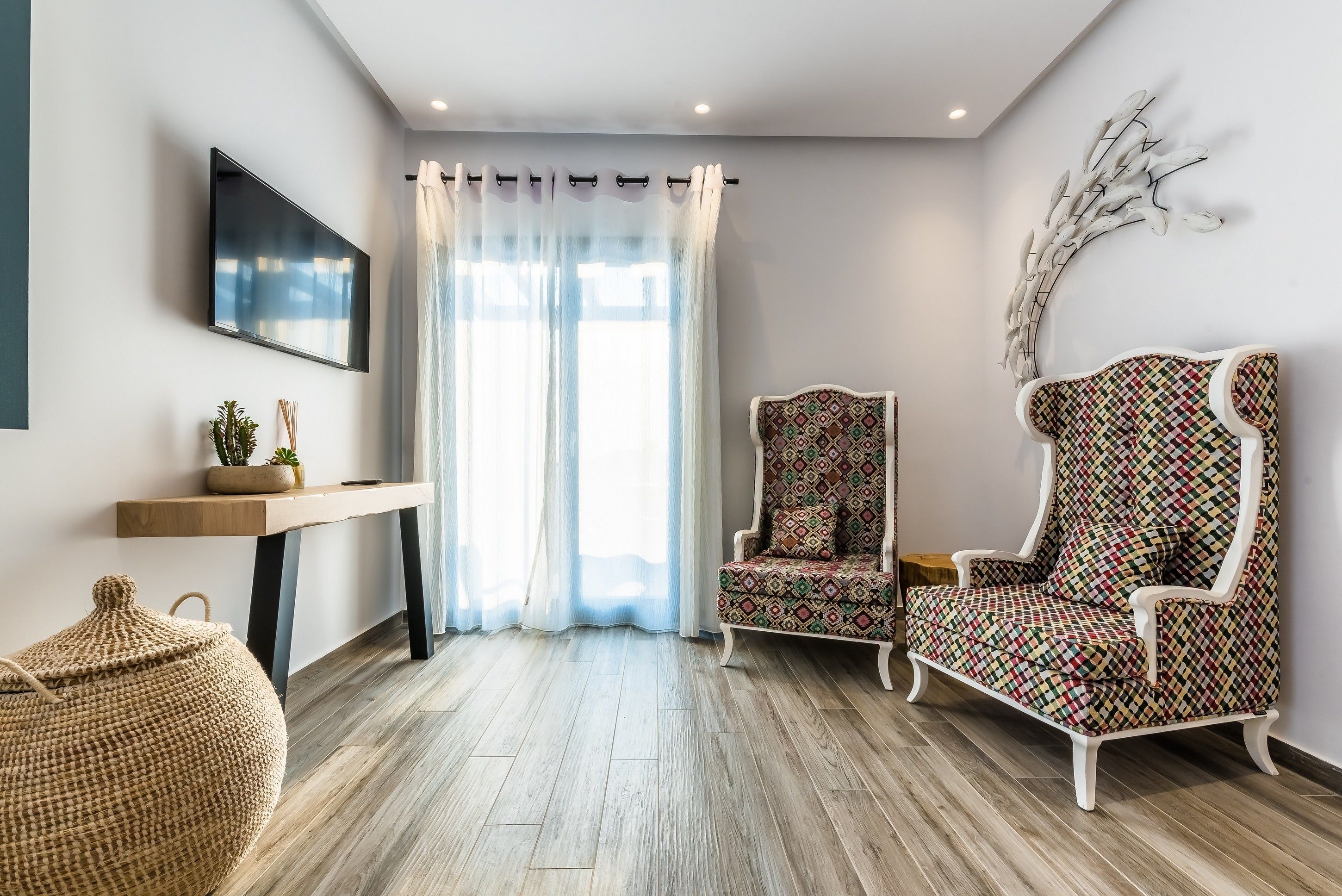 Ein modernes Wohnzimmer mit Holzboden, einem an der Wand montierten Fernseher, einem gemusterten Sessel, einem kleinen Tisch mit einer Pflanze und einem dekorativen Wandbild.