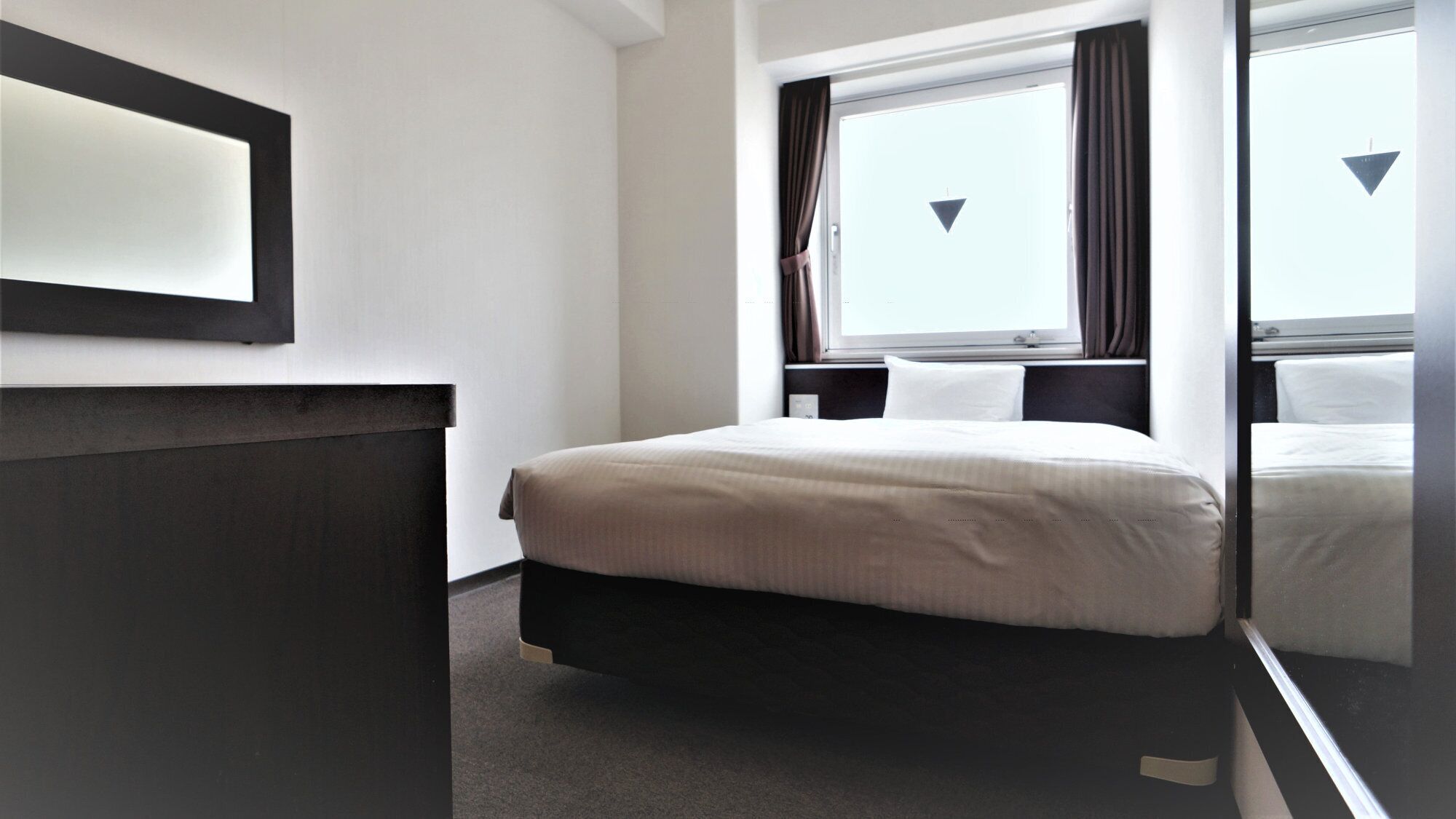 Una habitación de hotel con cama, mesita de noche, ventana con cortinas y un espejo montado en la pared.