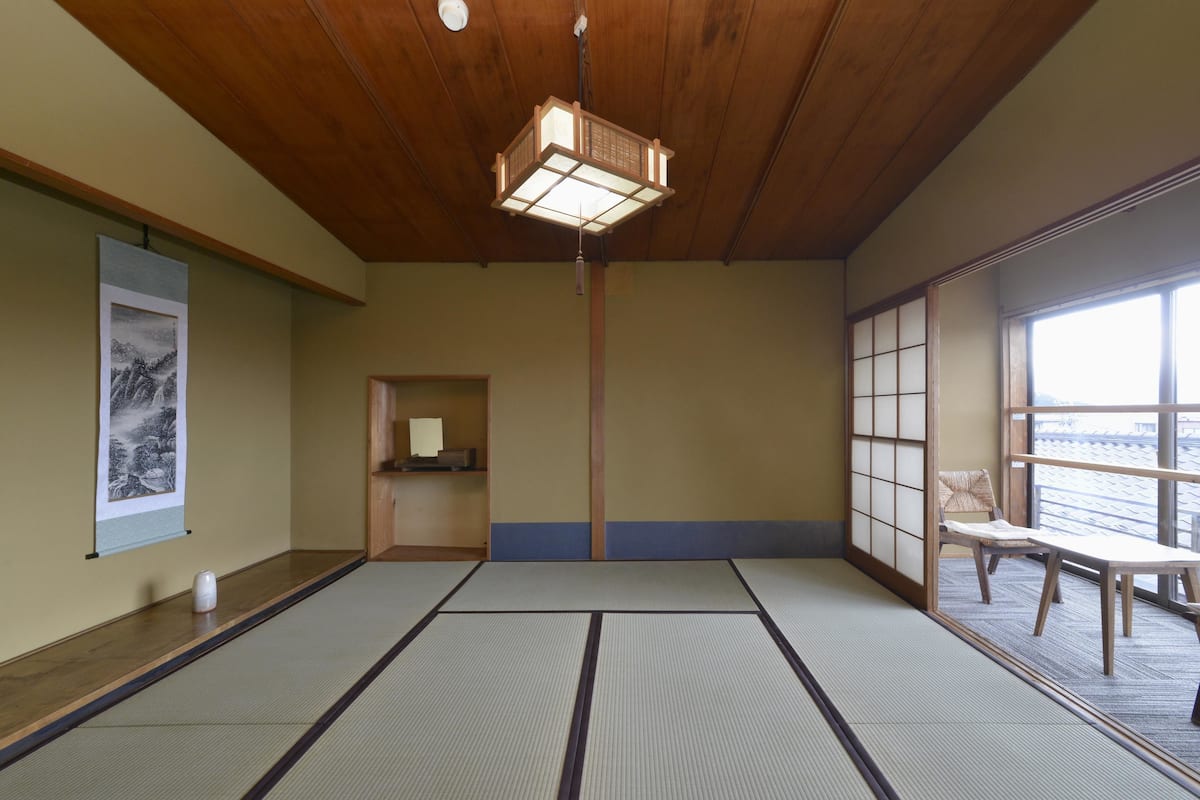 Una habitación con esteras de tatami, techo de madera, lámpara colgante, un cuadro en la pared, una mesita y una silla.