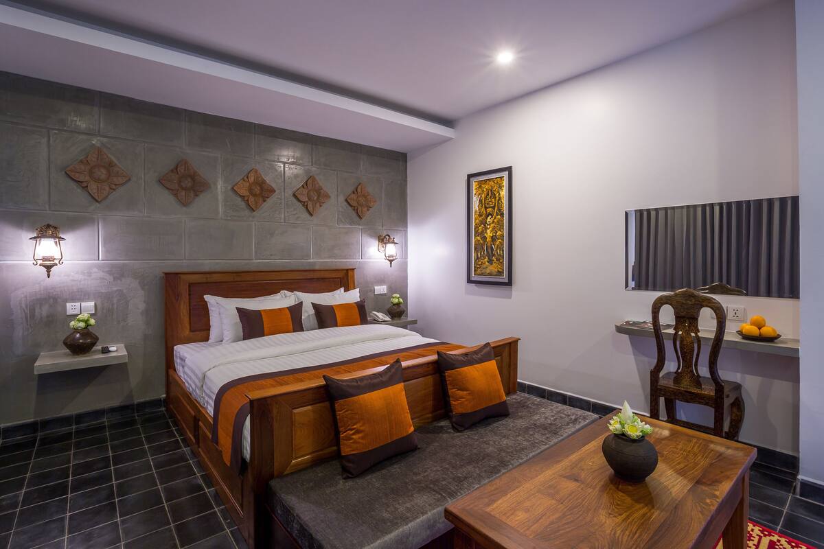 A neatly arranged hotel room with a large bed, bedside tables, a wooden bench, a small table with a vase, and a wall-mounted TV.