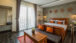A hotel room with a bed, a wooden table, a bench, a window with curtains, a wall-mounted light, and a small shelf with a vase. - Silver Palms Residence (Siem Reap)