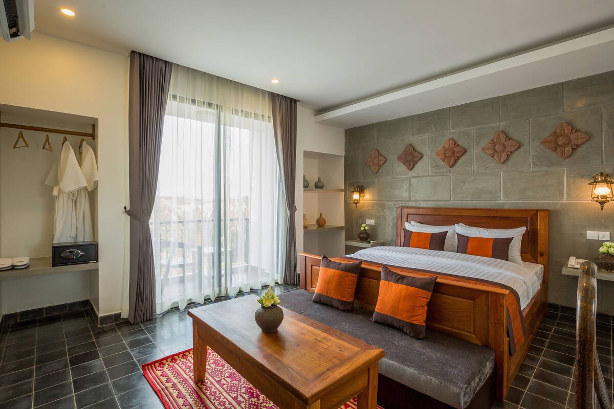 A hotel room with a bed, a wooden table, a bench, a window with curtains, a wall-mounted light, and a small shelf with a vase.