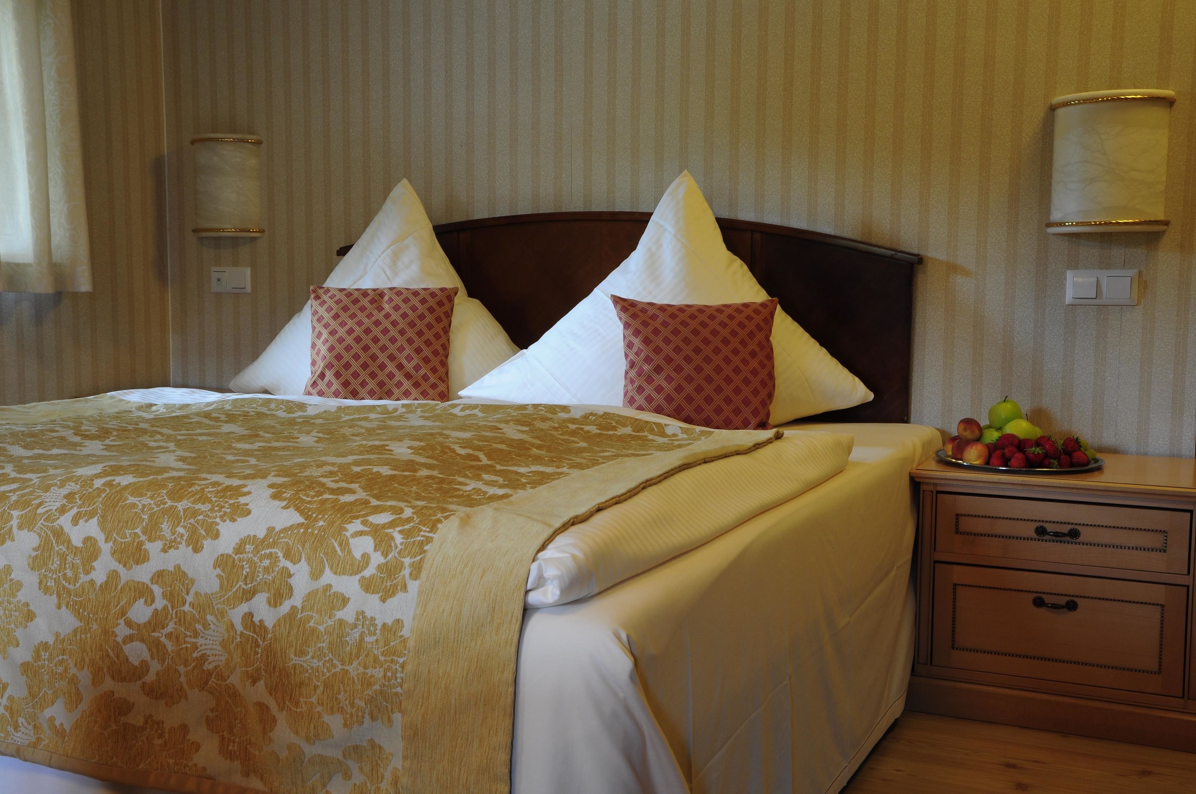 A neatly made bed with a patterned bedspread, two pillows with a diamond pattern, and a bedside table with a fruit bowl.