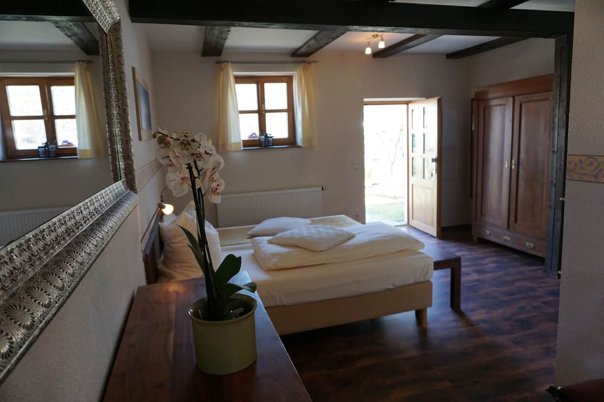 Ein Schlafzimmer mit einem Bett, einem Nachttisch mit einer Topf-Orchidee, einem großen Spiegel und einem Fenster mit Vorhängen.