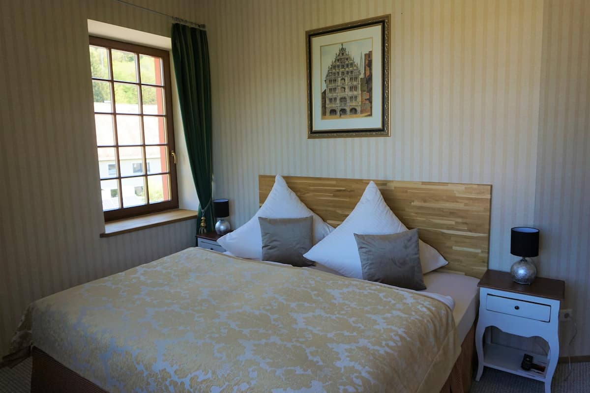 Ein ordentlich eingerichtetes Hotelzimmer mit einem großen Bett, einem Nachttisch, einer Fensterbank mit Vorhängen und einem gerahmten Bild an der Wand.