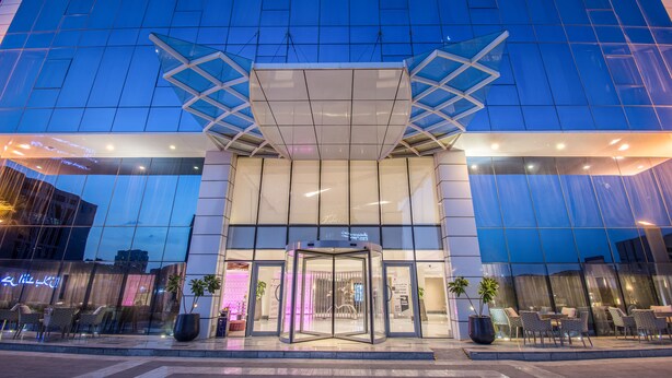 Una entrada de hotel moderna con puerta giratoria, fachada de vidrio y zona de estar al aire libre.