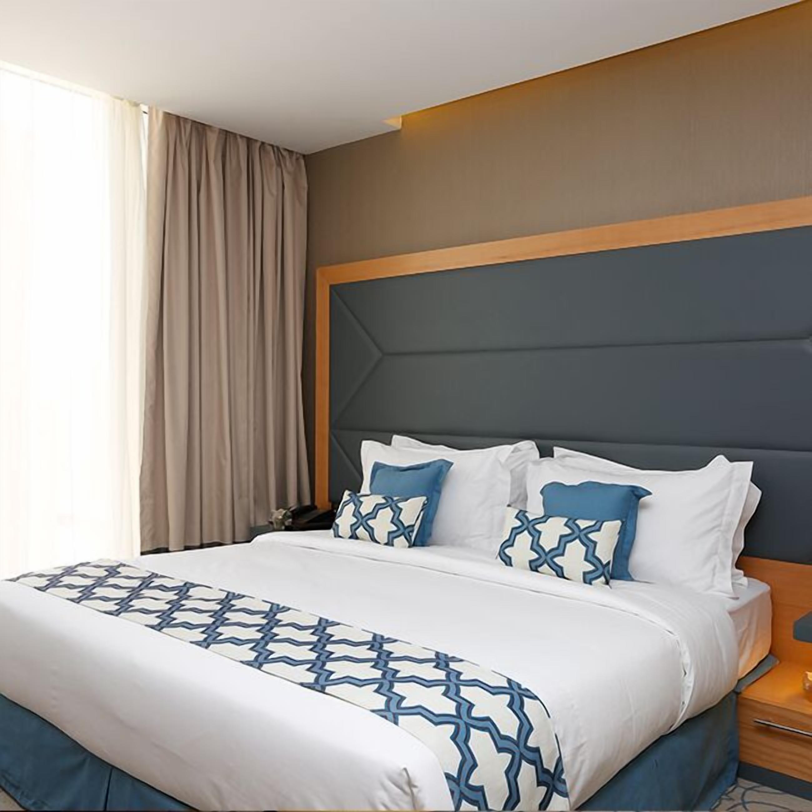 A hotel room with a large bed, a headboard with a geometric pattern, and a window with curtains.