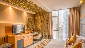 Una habitación de hotel moderna con una cama grande, un escritorio con computadora, una silla y vistas a la ciudad a través de amplios ventanales.