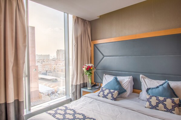 Habitación de hotel con un ventanal grande, una cama con almohadas estampadas, un jarrón con flores y una mesita de noche con un teléfono.
