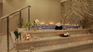 Un área de spa con una escalera de mármol, una bañera de hidromasaje y elementos decorativos como flores y velas.