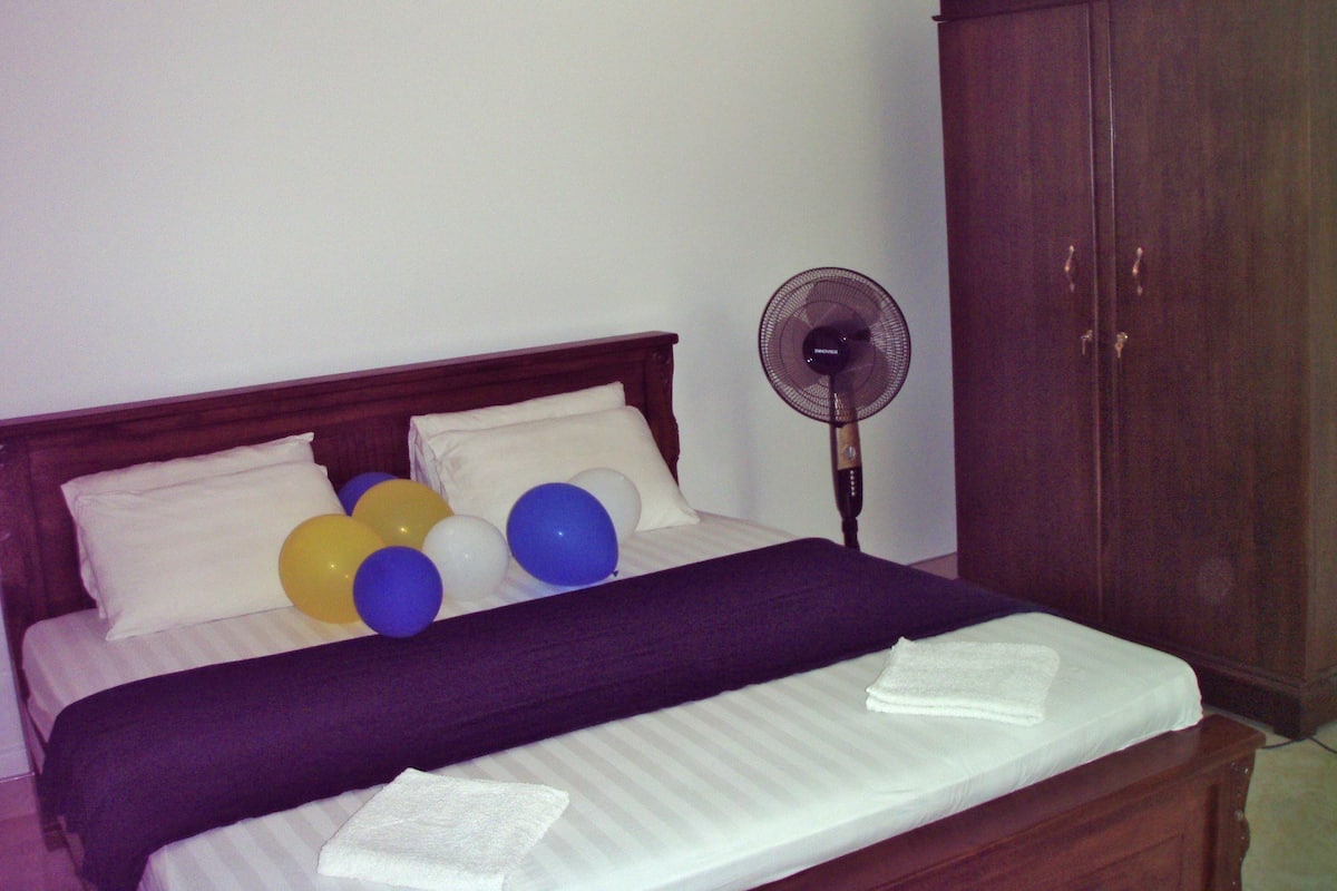 A bed with white linens, a dark blanket, and several balloons in blue, yellow, and white. A wooden wardrobe is to the right of the bed.