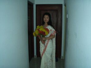 A person in a white saree holding a bouquet of flowers, standing in a hallway with a wooden door in the background.