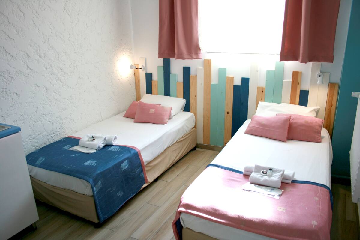 A hotel room with two beds, each with a different colored bedspread, a wooden headboard, and a window with curtains.