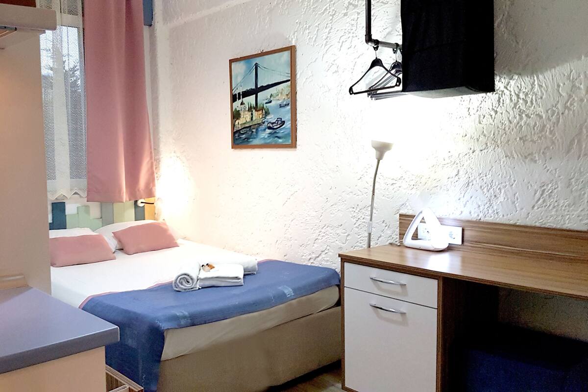 A bedroom with a bed, bedside table, desk, and a painting on the wall.