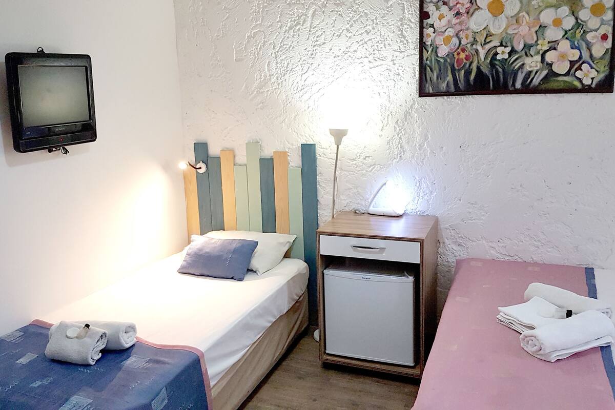 A small, neatly arranged room with a bed, a bedside table with a lamp, a small fridge, and a painting on the wall.