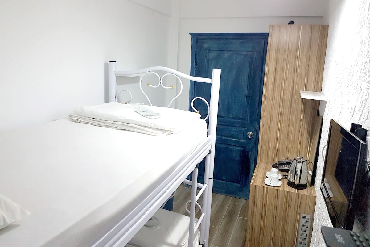 A small, single-bed room with a white metal bed frame, a wooden wardrobe, and a small table with cups and a kettle.