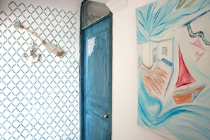 Une porte bleue ornée d’un motif décoratif, une fenêtre avec un grillage métallique et un tableau représentant des voiliers sur le mur.