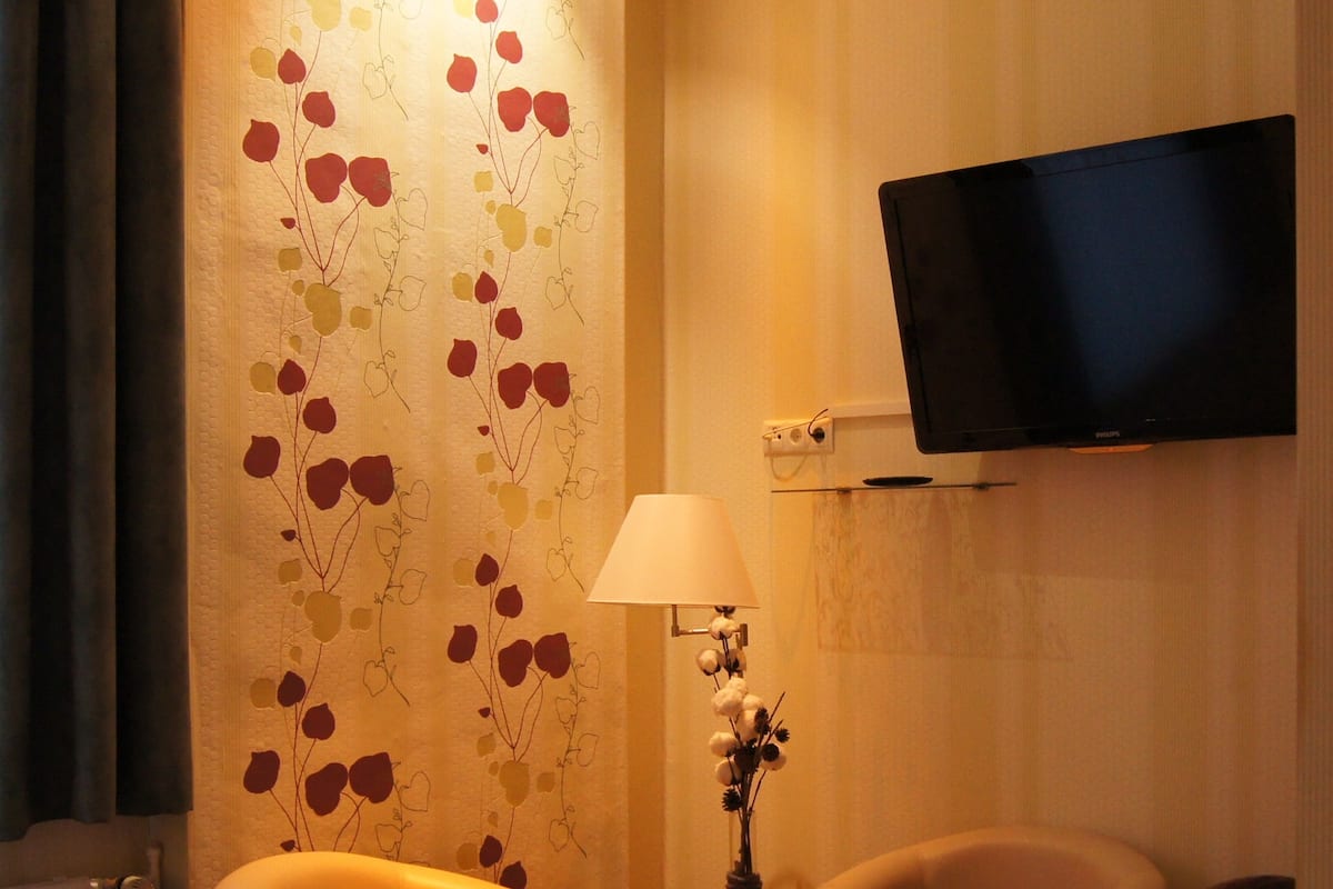 Hotellihuone, jossa on sänky, kaksi nojatuolia, pieni pöytä lampun kanssa, maljakko kukkineen, seinälle asennettu televisio ja kukkakuviollinen tapetti.