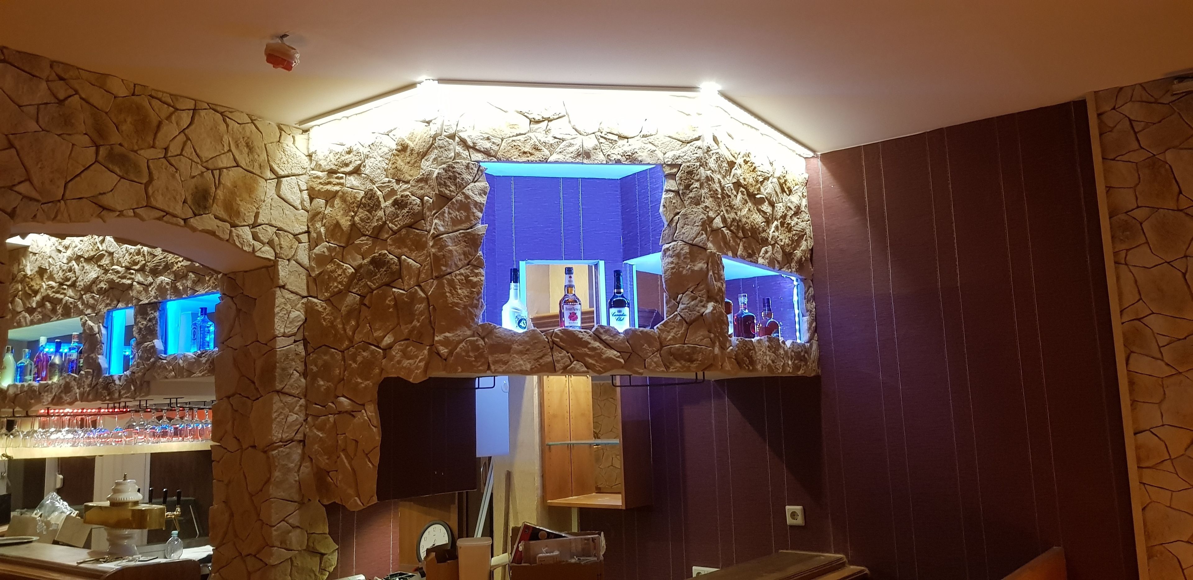 Zona de bar con paredes de piedra, iluminación azul y diferentes botellas y vasos.