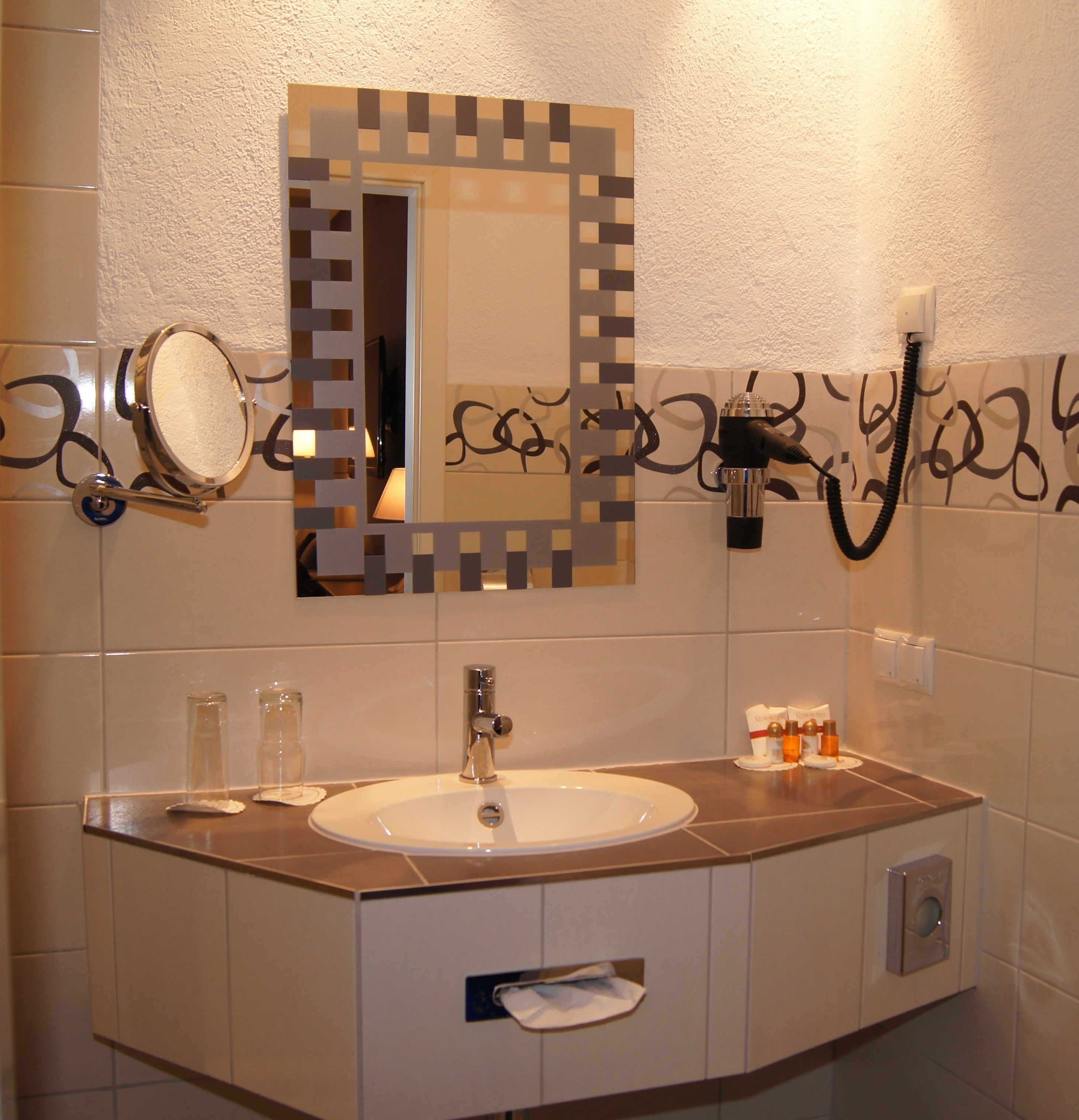 A bathroom with a white sink, mirror, and vanity. There are two glass bottles on the counter, a hairdryer, and a towel dispenser.