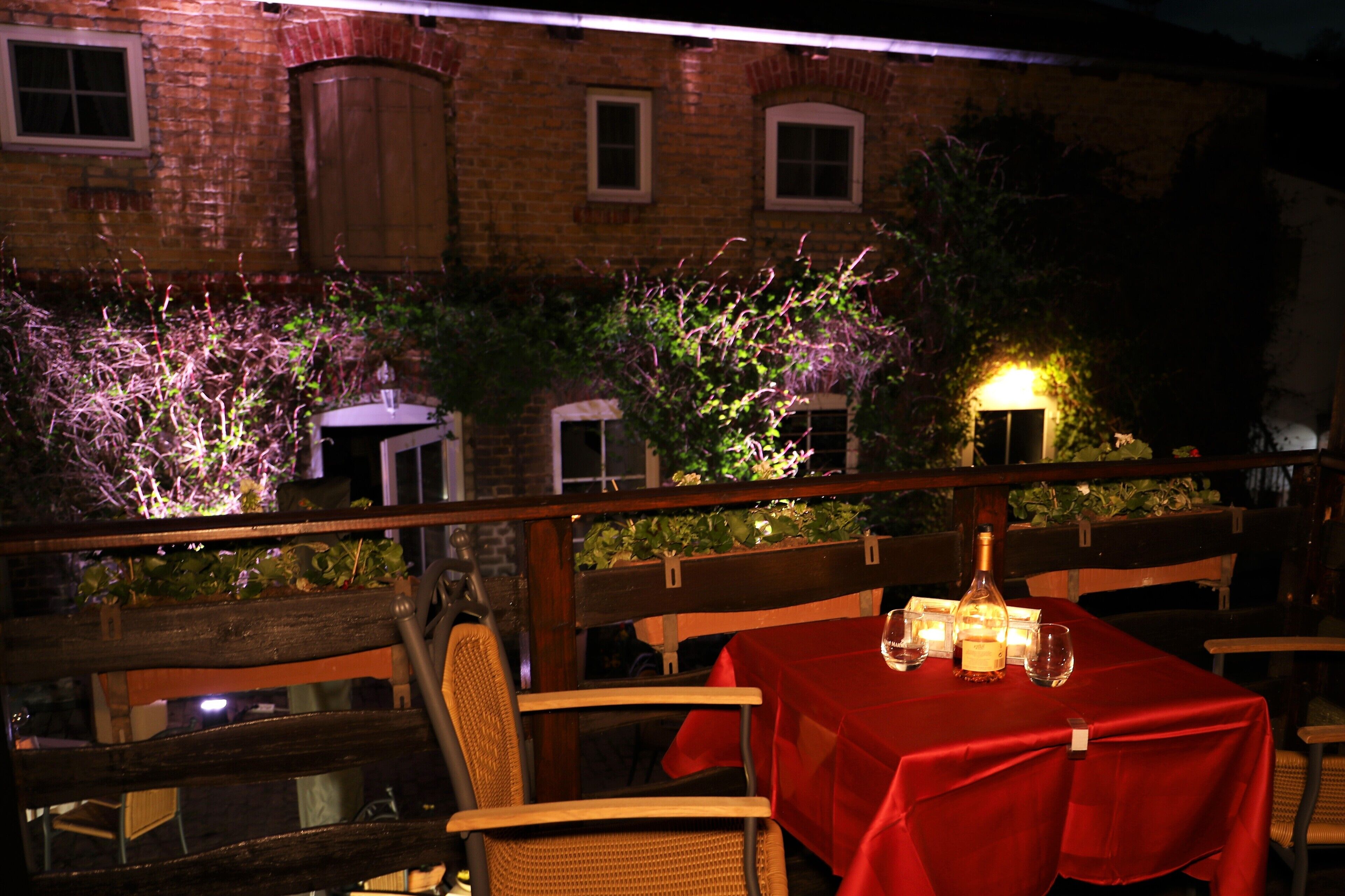 Un comedor al aire libre con mantel rojo, sillas de mimbre y una botella de vino.