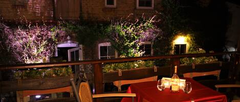 Un comedor al aire libre con mantel rojo, sillas de mimbre y una botella de vino.