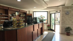 Un vestíbulo de hotel con mostrador de recepción, estantes con diversos objetos, un reloj y vistas al exterior.