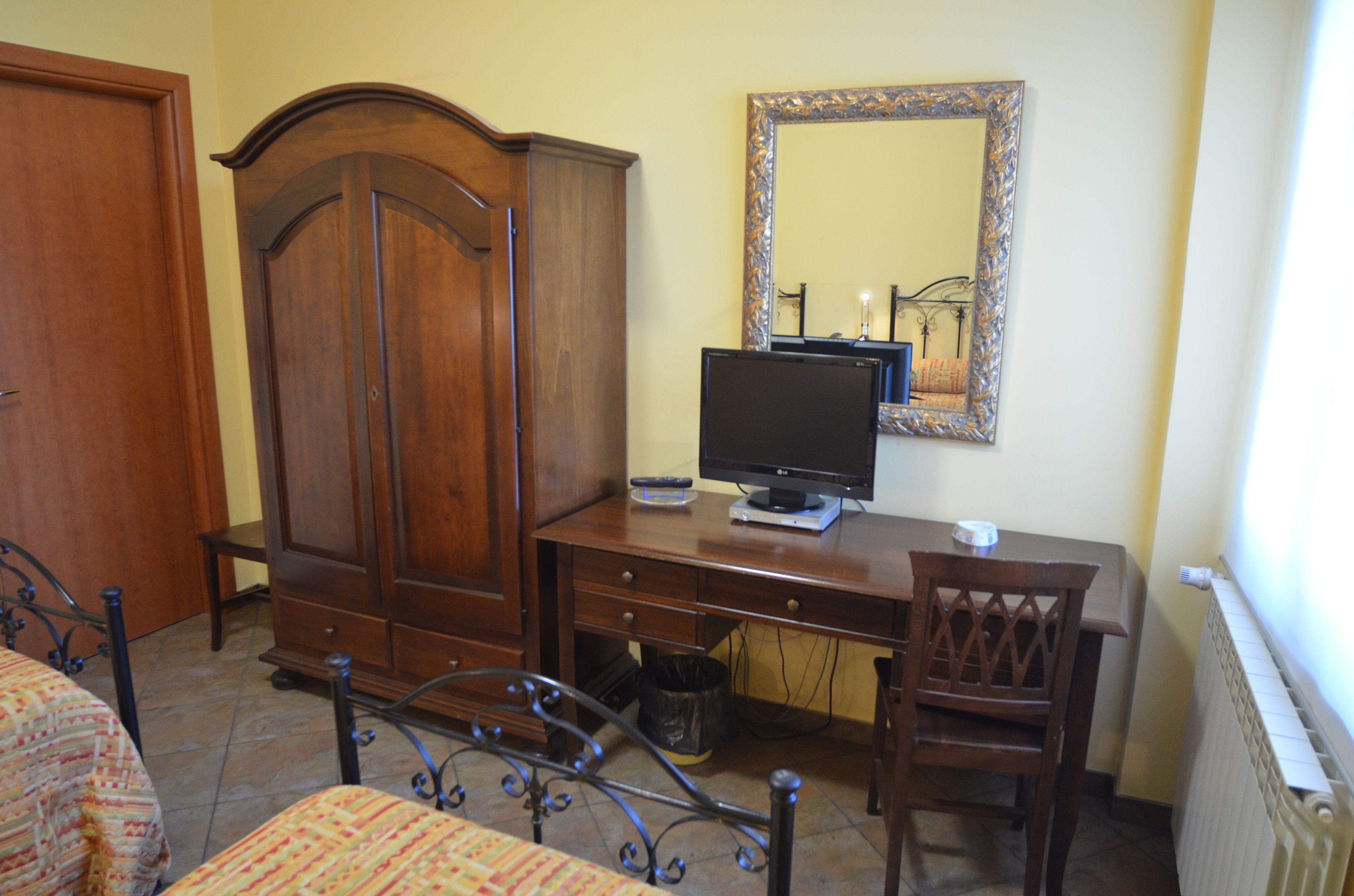 A bedroom with a wooden wardrobe, a desk with a computer and monitor, a chair, and a bed with patterned bedding.