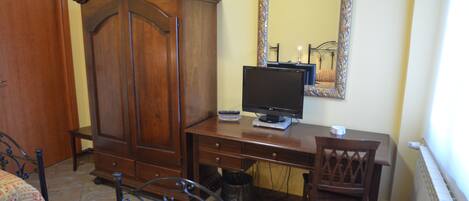 A bedroom with a wooden wardrobe, a desk with a computer and monitor, a chair, and a bed with patterned bedding.