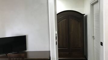 A room with a dark wooden door, a white wall, a dark wooden desk with a chair, and a television.