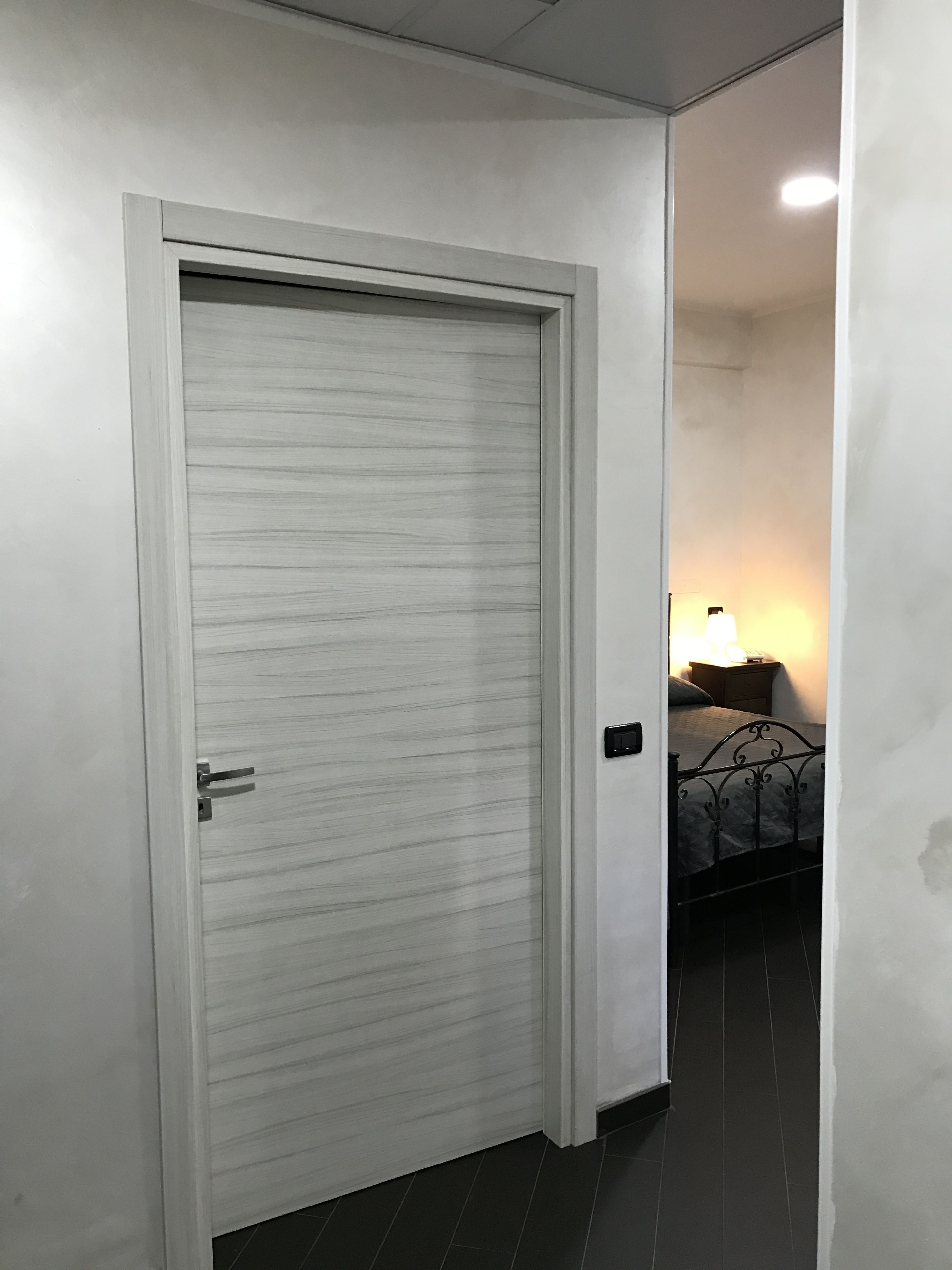 Una puerta blanca cerrada con picaporte, un suelo de baldosas oscuras y una cama con estructura metálica y ropa de cama blanca en la habitación contigua.