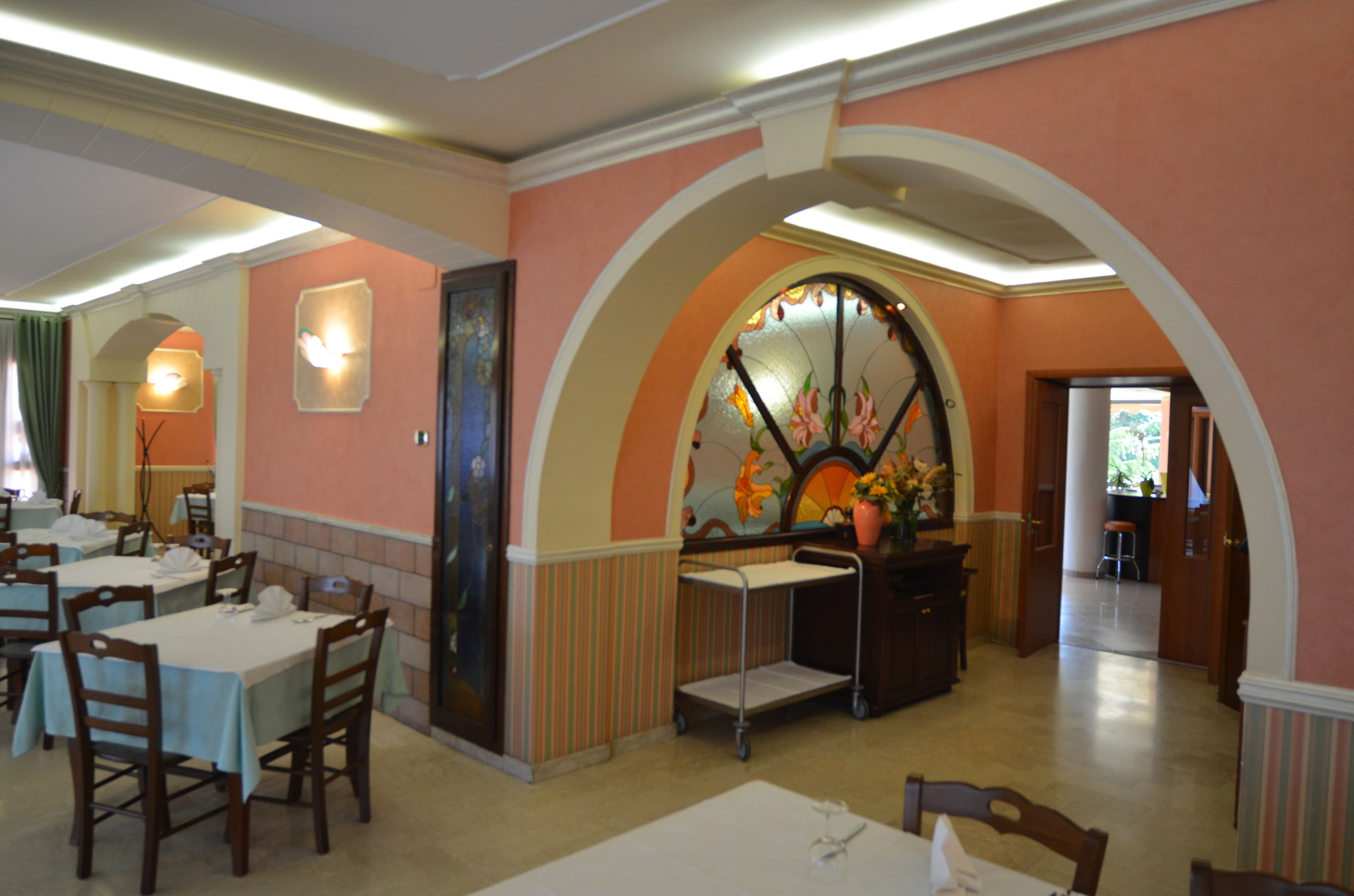 Un comedor con mesas y sillas, un muro decorativo con diseño floral, y un pasillo que conduce a otra habitación.