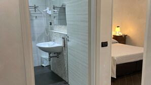 Un baño moderno con lavamanos, espejo y ducha. En la habitación contigua, hay una cama con mesita de noche y lámpara.