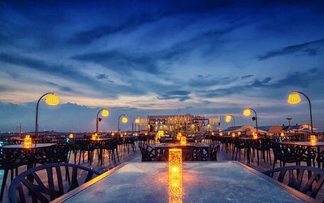 An outdoor dining area with tables and chairs, street lamps, and a view of the cityscape at dusk.