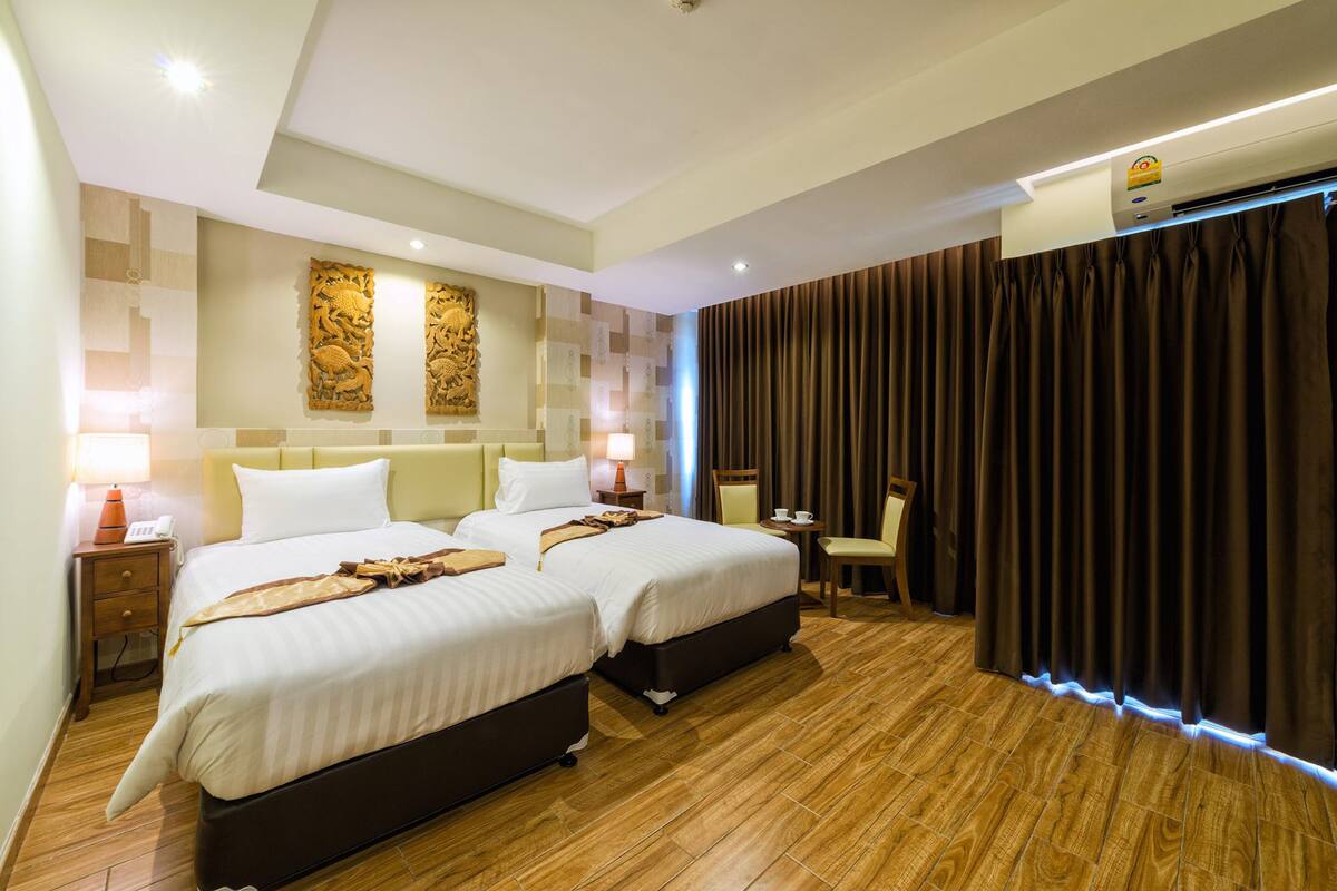 A hotel room with two beds, a small table, a chair, a nightstand, and a wall-mounted air conditioner.