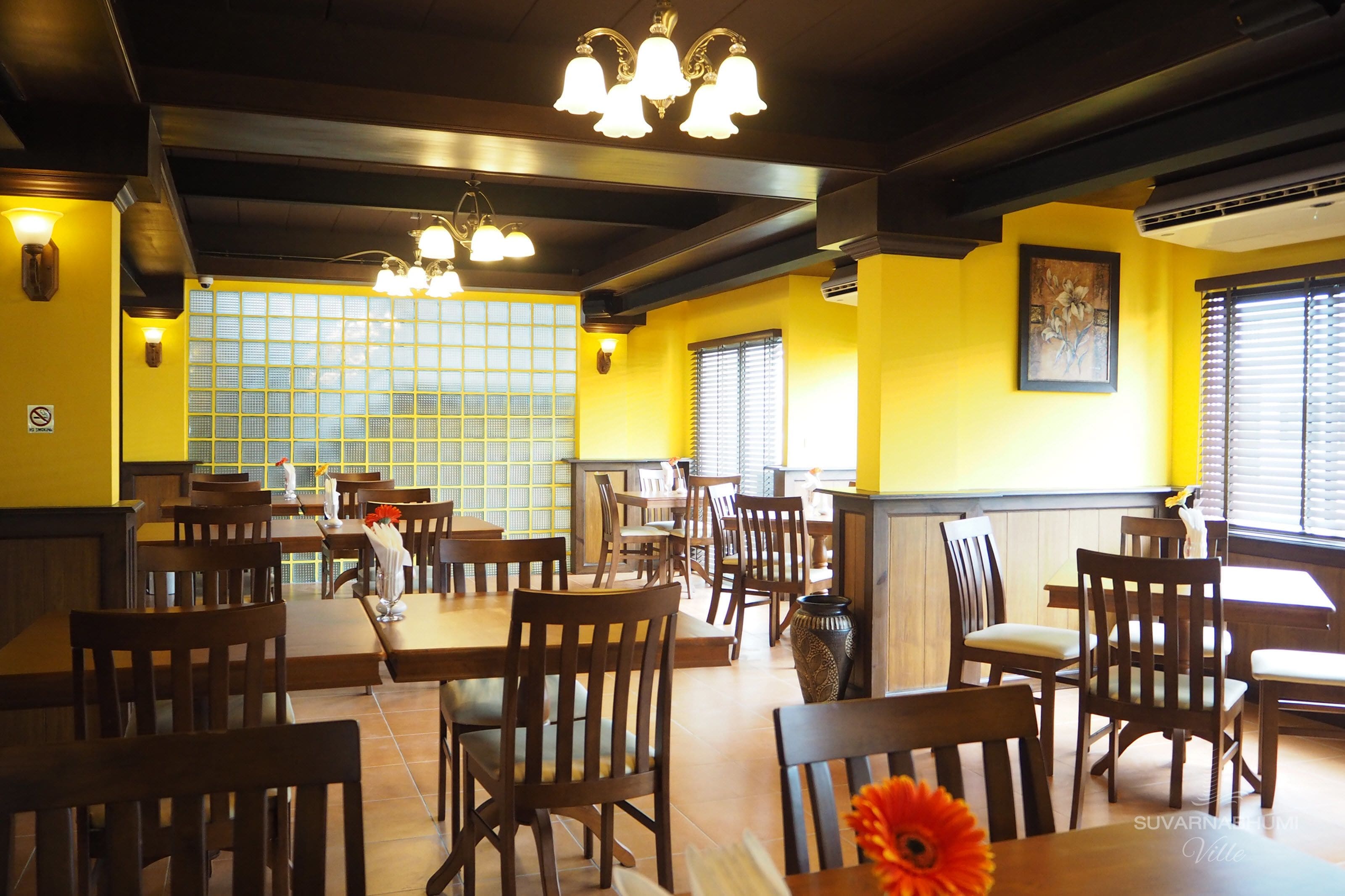 A restaurant with wooden tables and chairs, yellow walls, and a large window with blinds.