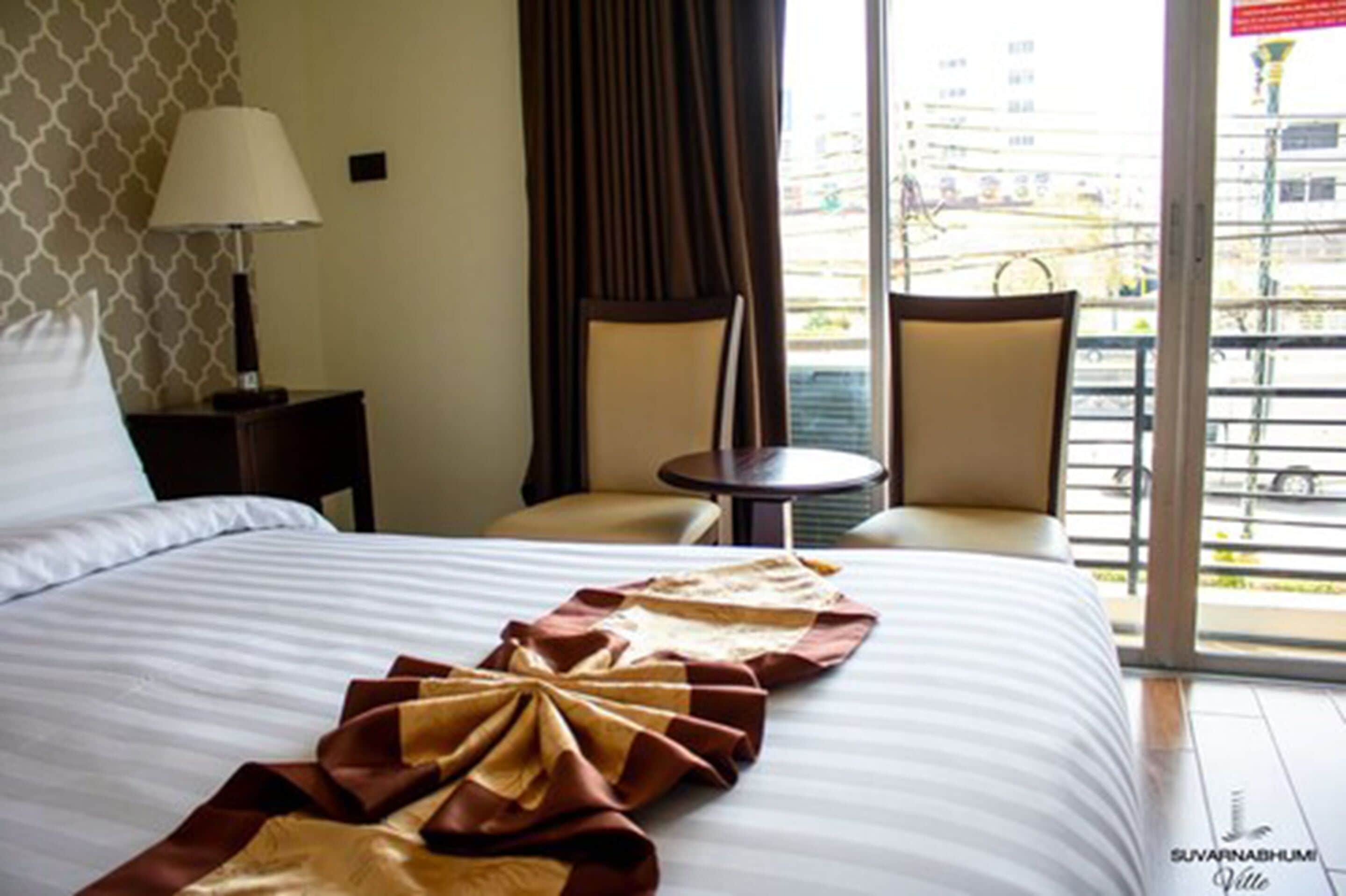 A hotel room with a bed, two chairs, a small table, a lamp, and a view of the outside.