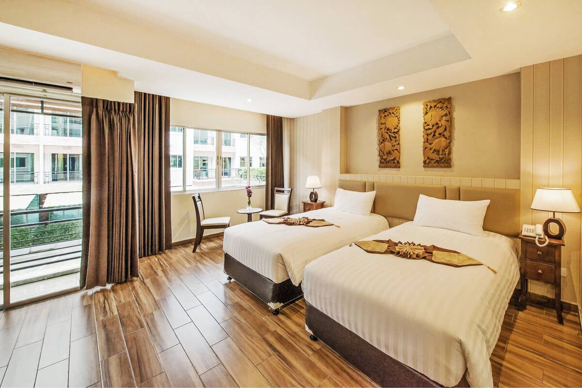 A hotel room with two beds, a sitting area with a chair and table, and large windows offering a view of the building outside.