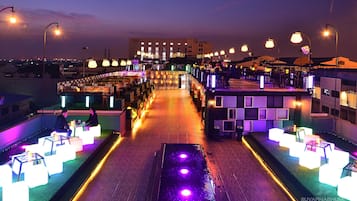 A rooftop bar with illuminated tables and chairs, a pool, and a view of the city skyline at dusk.
