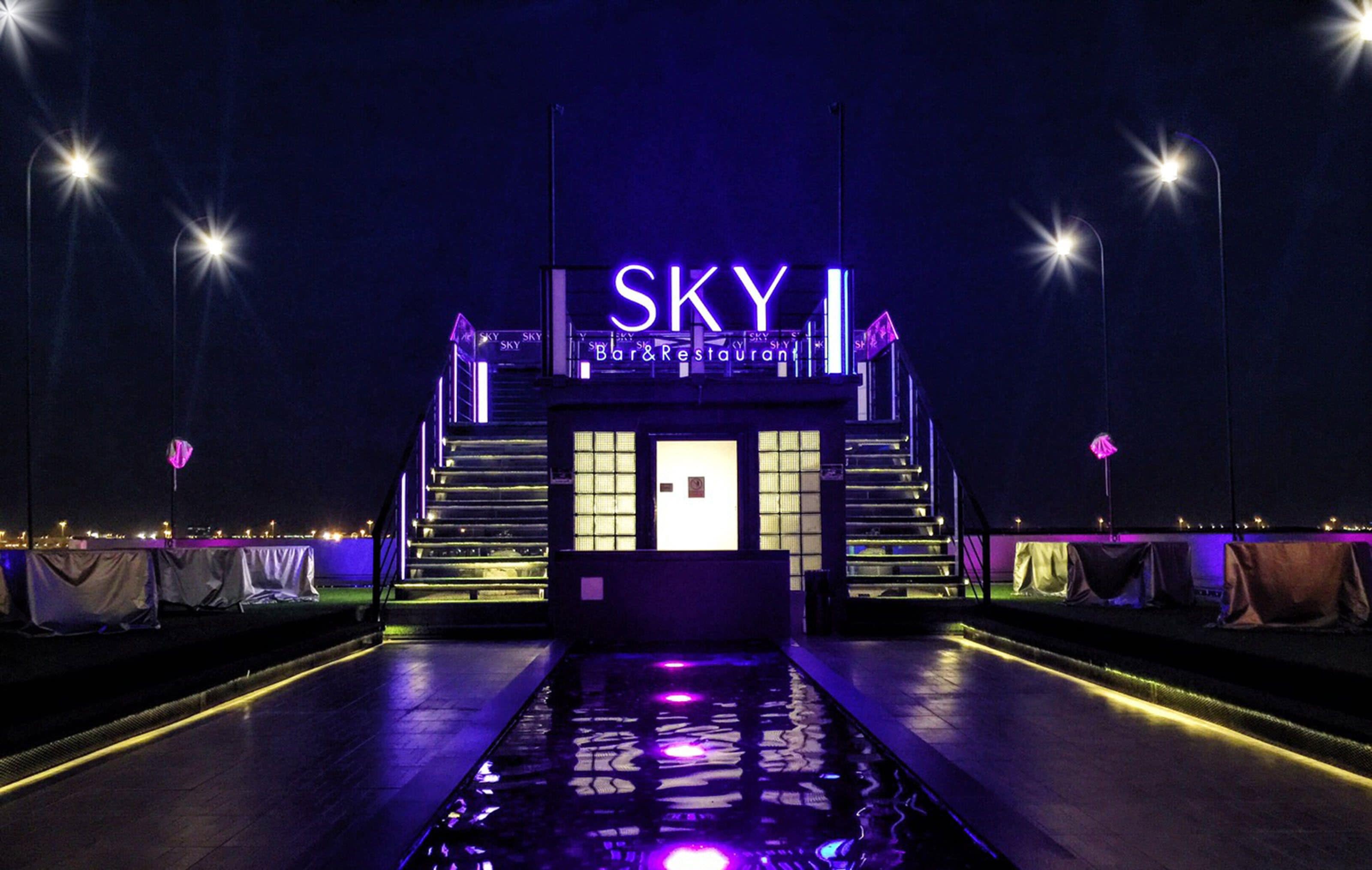 A nighttime view of the SKY Bar & Restaurant with illuminated signage and a reflective water feature in the foreground.