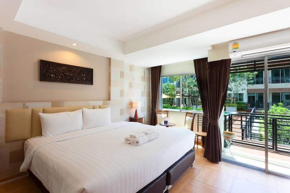 A hotel room with a large bed, a view of the outdoors, a small table with a chair, and a balcony with a railing.
