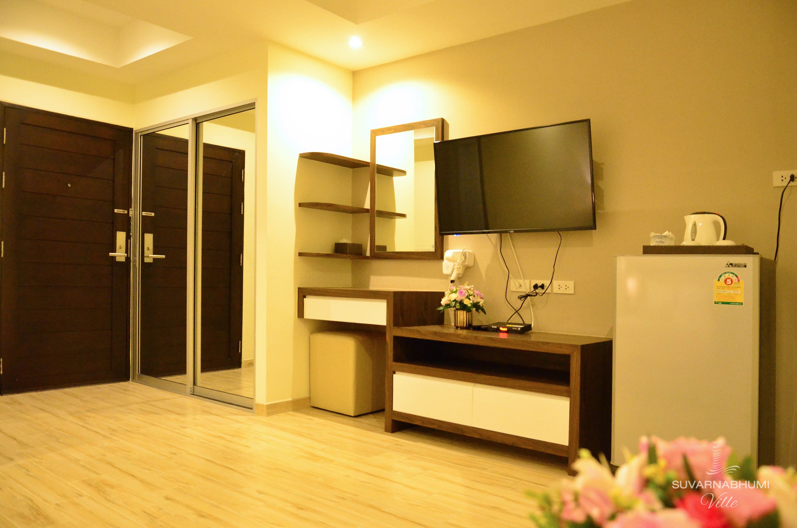 A hotel room with a wooden floor, a sliding door, a flat-screen TV mounted on the wall, a small refrigerator, and a wooden cabinet with a mirror.