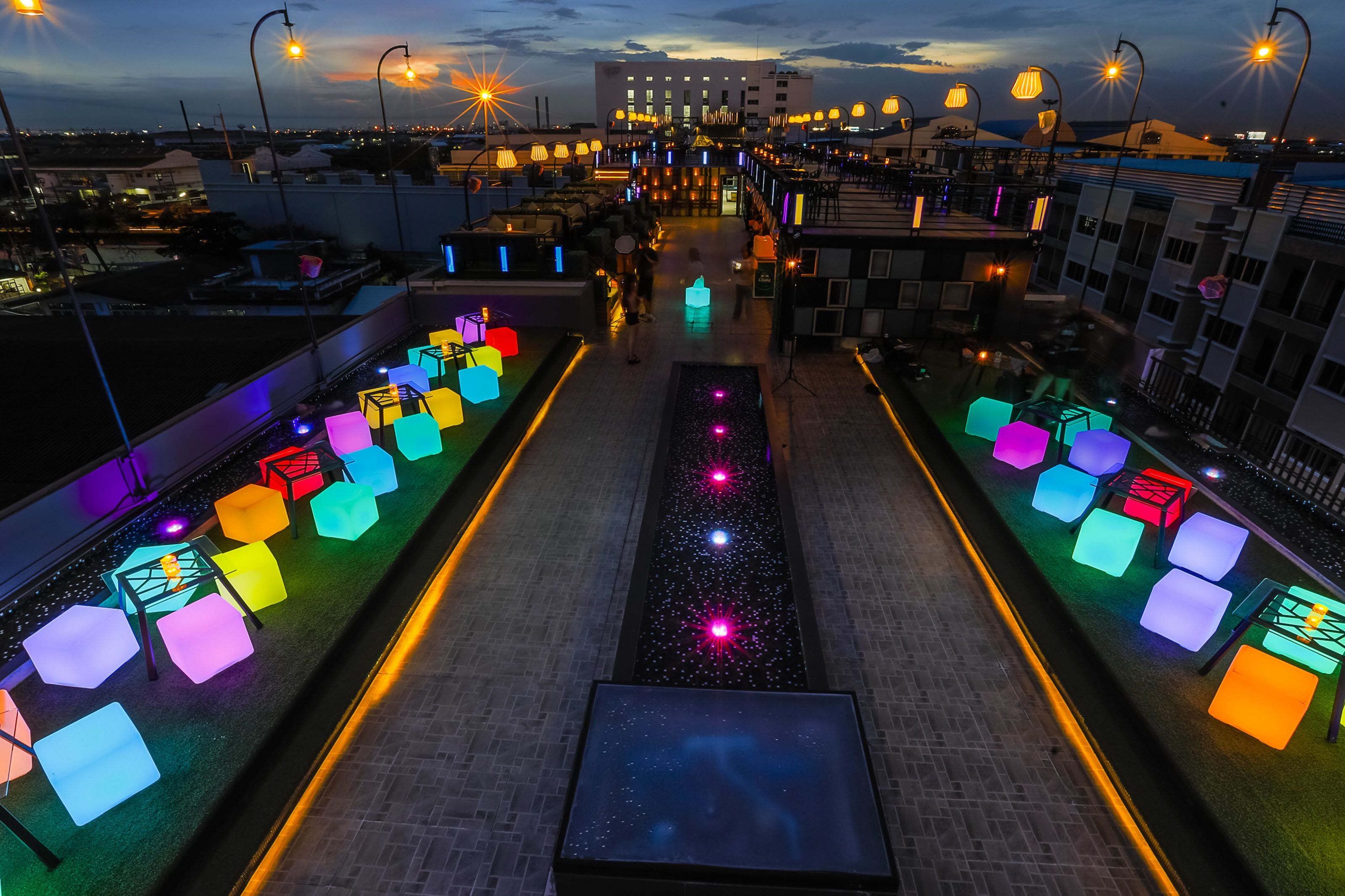 Una terraza en la azotea con zonas de estar iluminadas de colores y un paseo central.