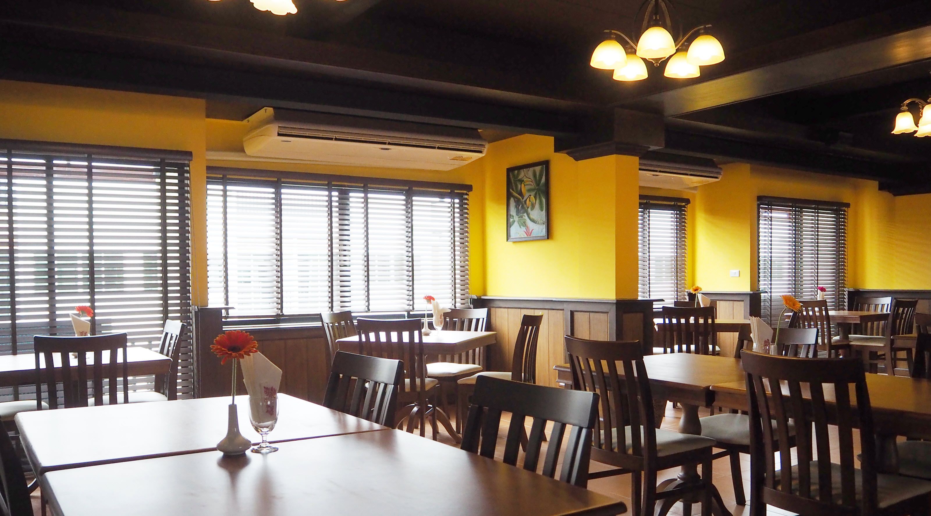 A restaurant interior with multiple tables and chairs, yellow walls, and large windows with blinds.
