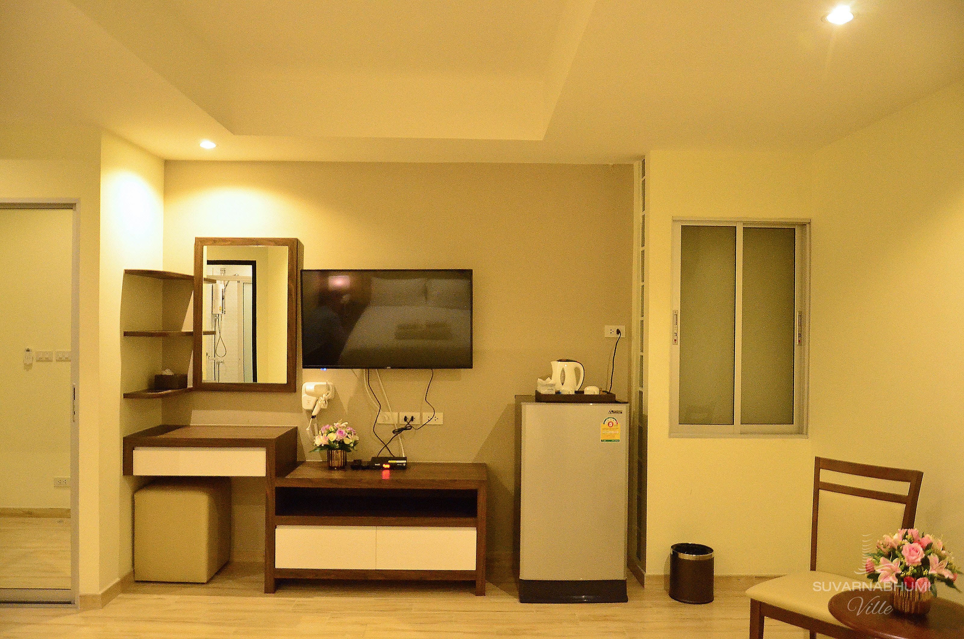 A compact living space with a television mounted on the wall, a small refrigerator, a wooden cabinet, and a chair with a flower vase.