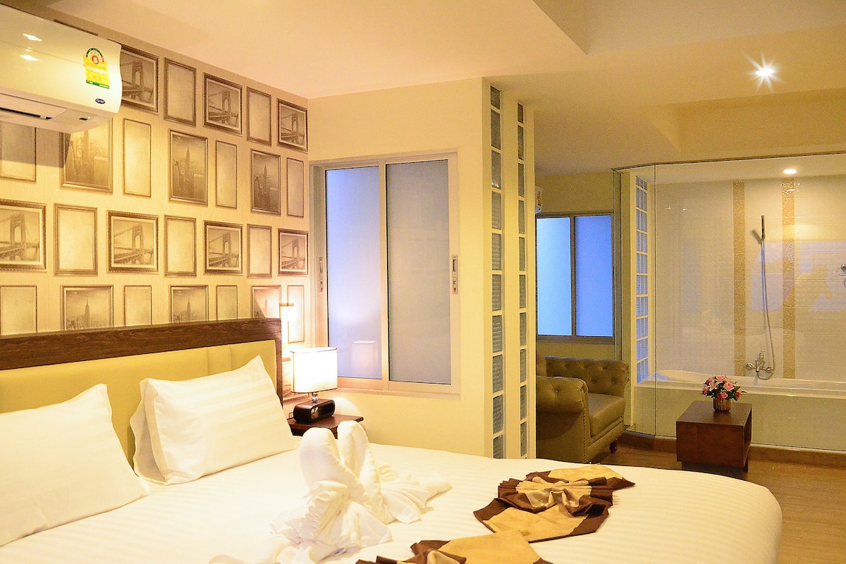 A hotel room with a bed, a nightstand, a lamp, a bathroom with a shower, and a wall with framed pictures.