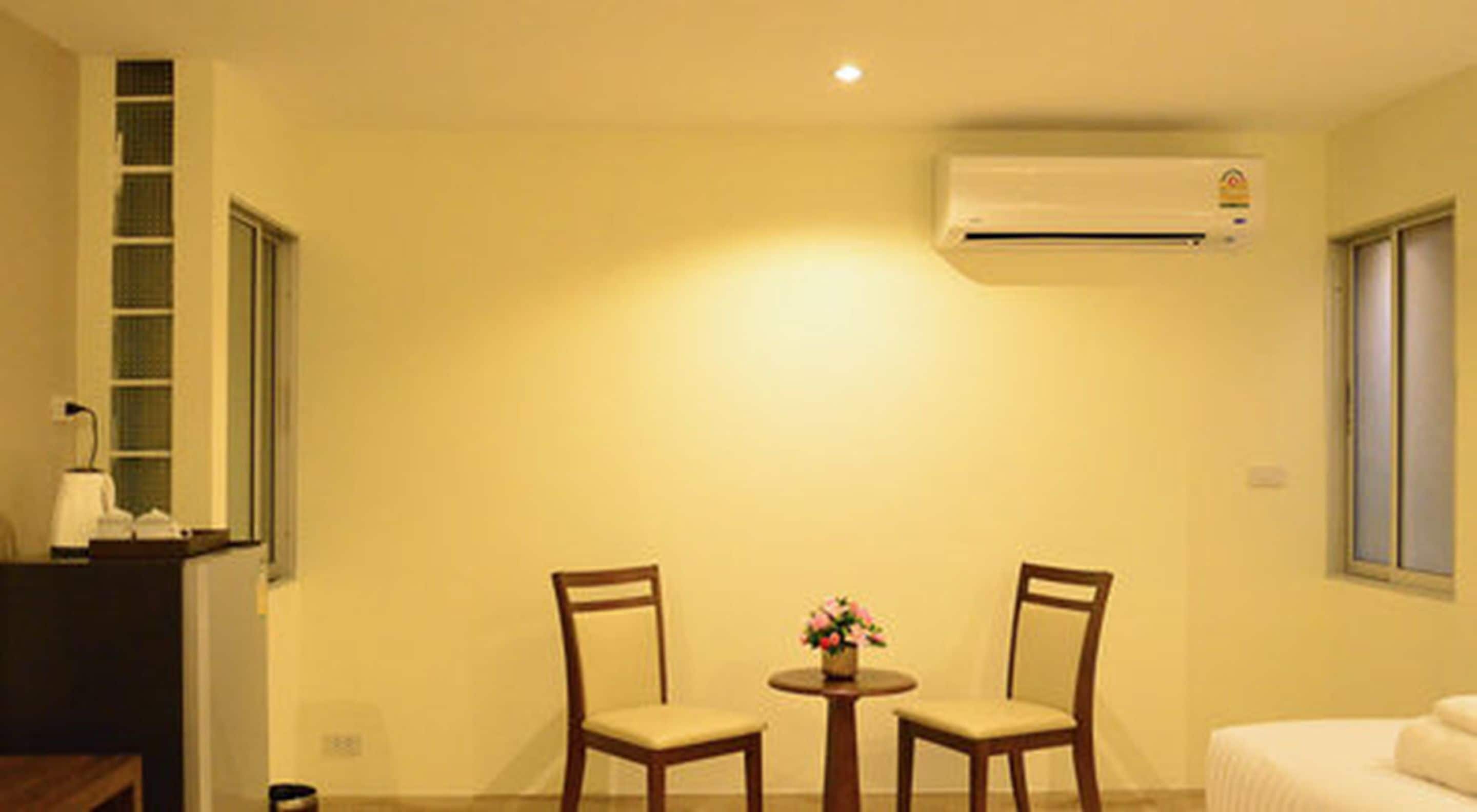 A small, well-lit room with two wooden chairs, a small table with a flower vase, an air conditioning unit on the wall, and a window with blinds.