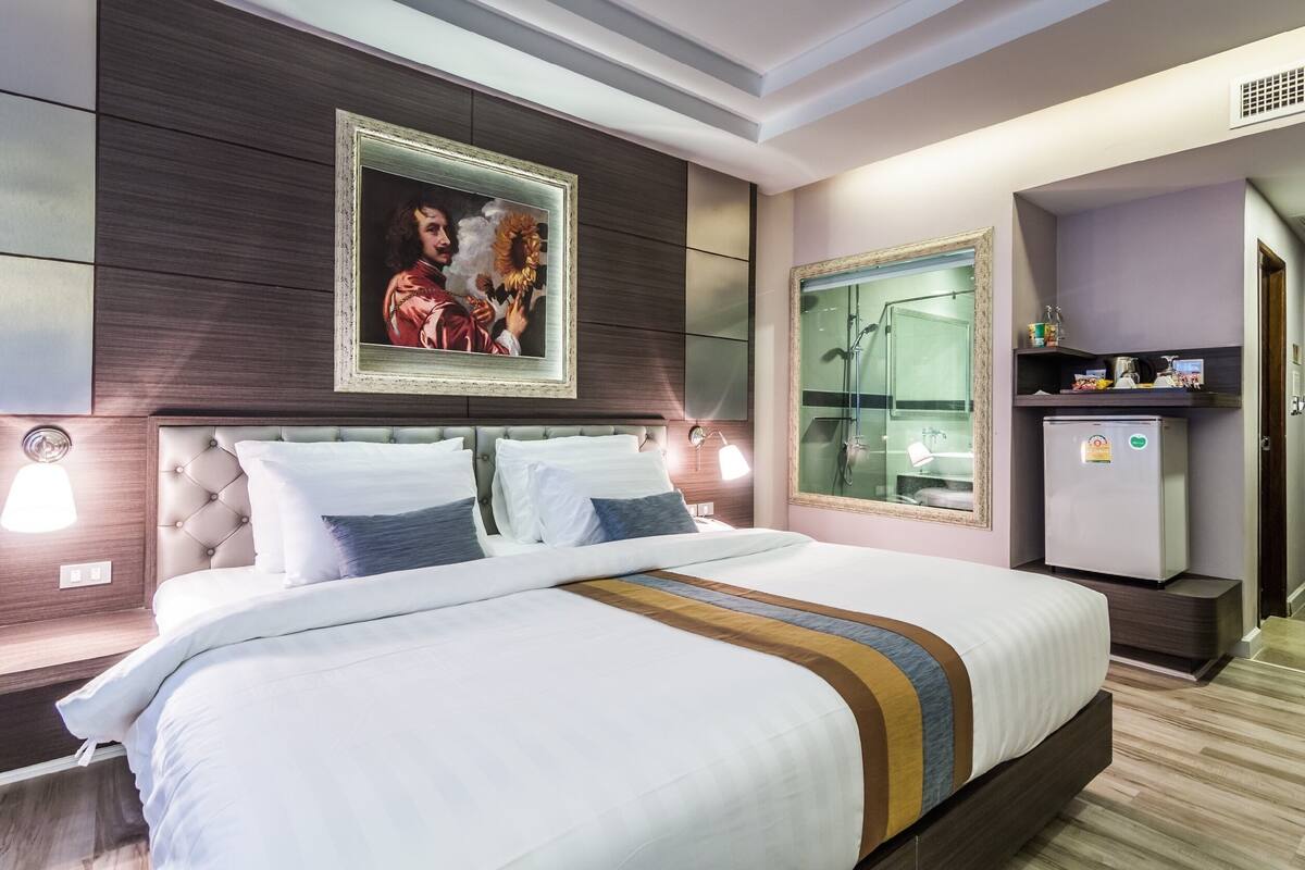 Una habitación de hotel moderna con una cama grande, un retrato enmarcado en la pared, una tocador con espejo y un refrigerador pequeño.