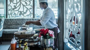 A chef preparing food in a well-lit, elegant dining area with decorative screens and a comfortable seating area in the background. - Gallery Design Hotel (Sisaket)