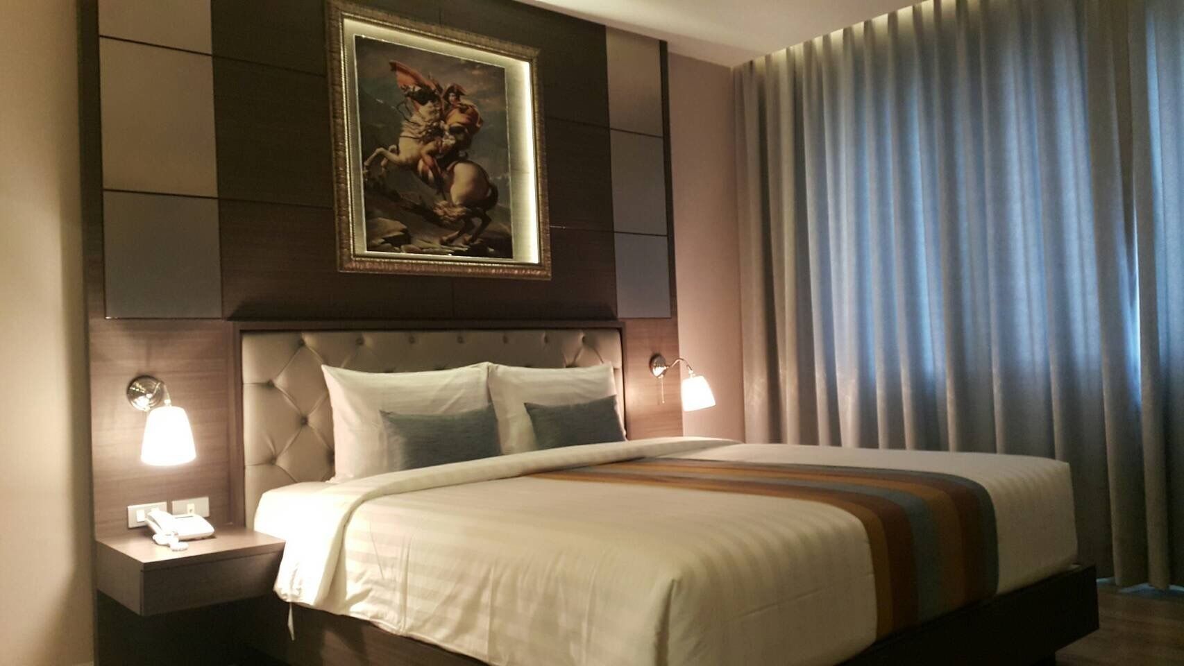 A hotel room with a large bed, a headboard with a painting, two bedside lamps, and a view of a curtain.